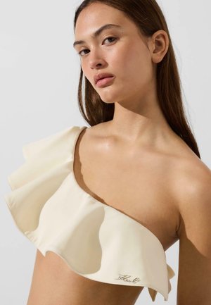 Tânără cu păr lung și maro, purtând un top crem pe umeri cu volane și un mic logo metalic în față.