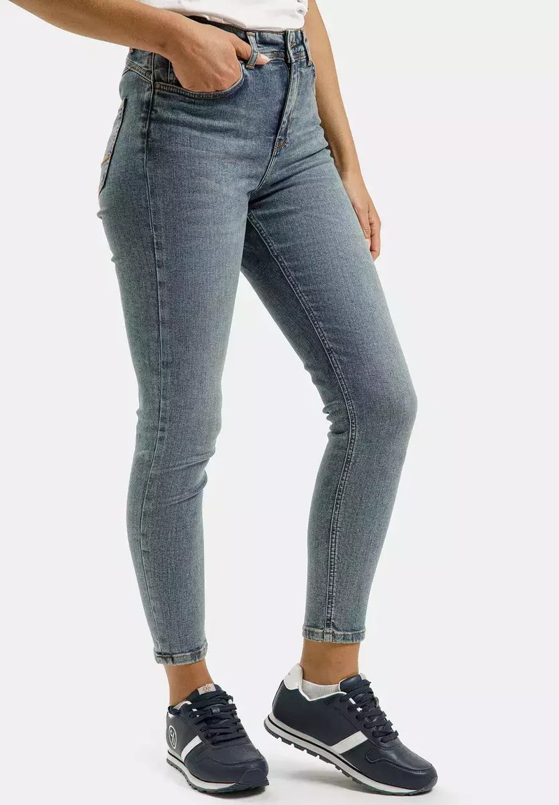 Jeans z wysokim stanem, dopasowane, w wyblakłym niebieskim denimi, z pięcioma kieszeniami i subtelnymi detalami w szyciu. W połączeniu z ciemnymi butami sportowymi.