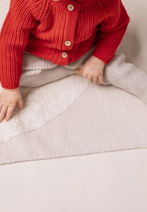 Małe dziecko ubrane w czerwony dziergany sweter i beżowe spodnie siedzi po turecku na warstwowej beżowej wykładzinie lub dywanie.