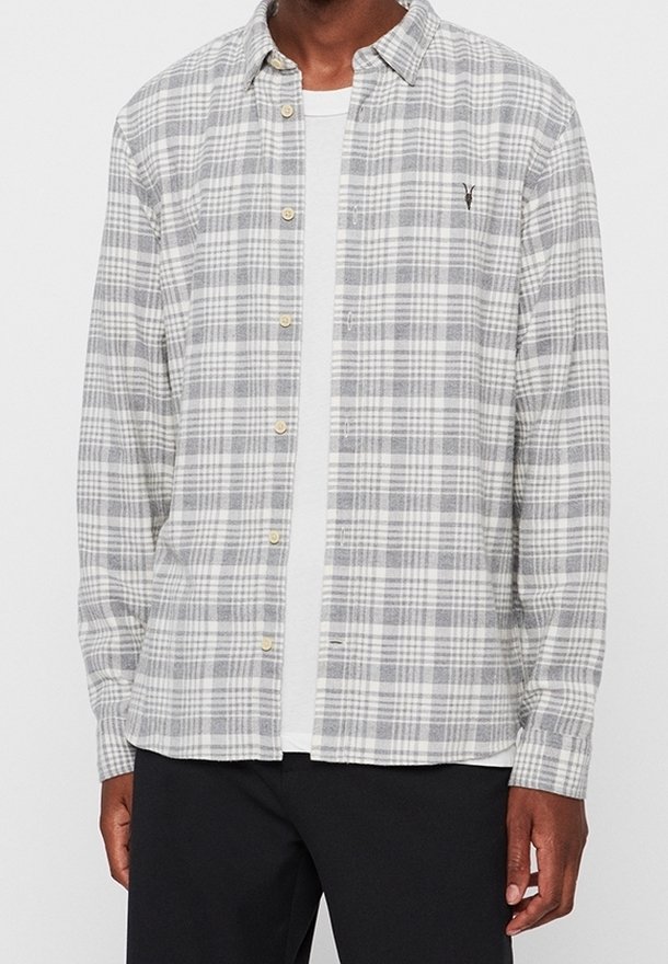 Homme portant une chemise boutonnée à carreaux grise et blanche sur un t-shirt blanc avec un pantalon noir, visible du cou jusqu'aux hanches.