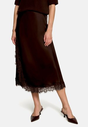Kobieta ubrana w ciemnobrązową spódnicę midi z koronkową lamówką i brązowe buty na obcasie typu slingback z zaostrzonym noskiem, stojąca na jednolitym tle.
