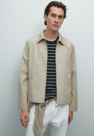 Beige lightweight jacket with a collared neck, side pockets, and a straight cut. Worn over a black and white striped shirt.