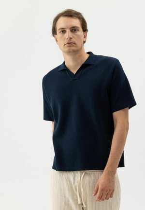 Világos bőrű, barna hajú férfi tengerészkék rövid ujjú pólóinget és világos bézs színű, húzózsinóros nadrágot visel, fehér háttér előtt áll.