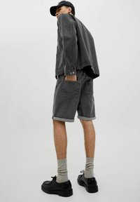 Veste en denim noir et shorts, avec des poignets roulés. La tenue présente une coupe décontractée et est associée à des chaussures noires et des chaussettes grises.
