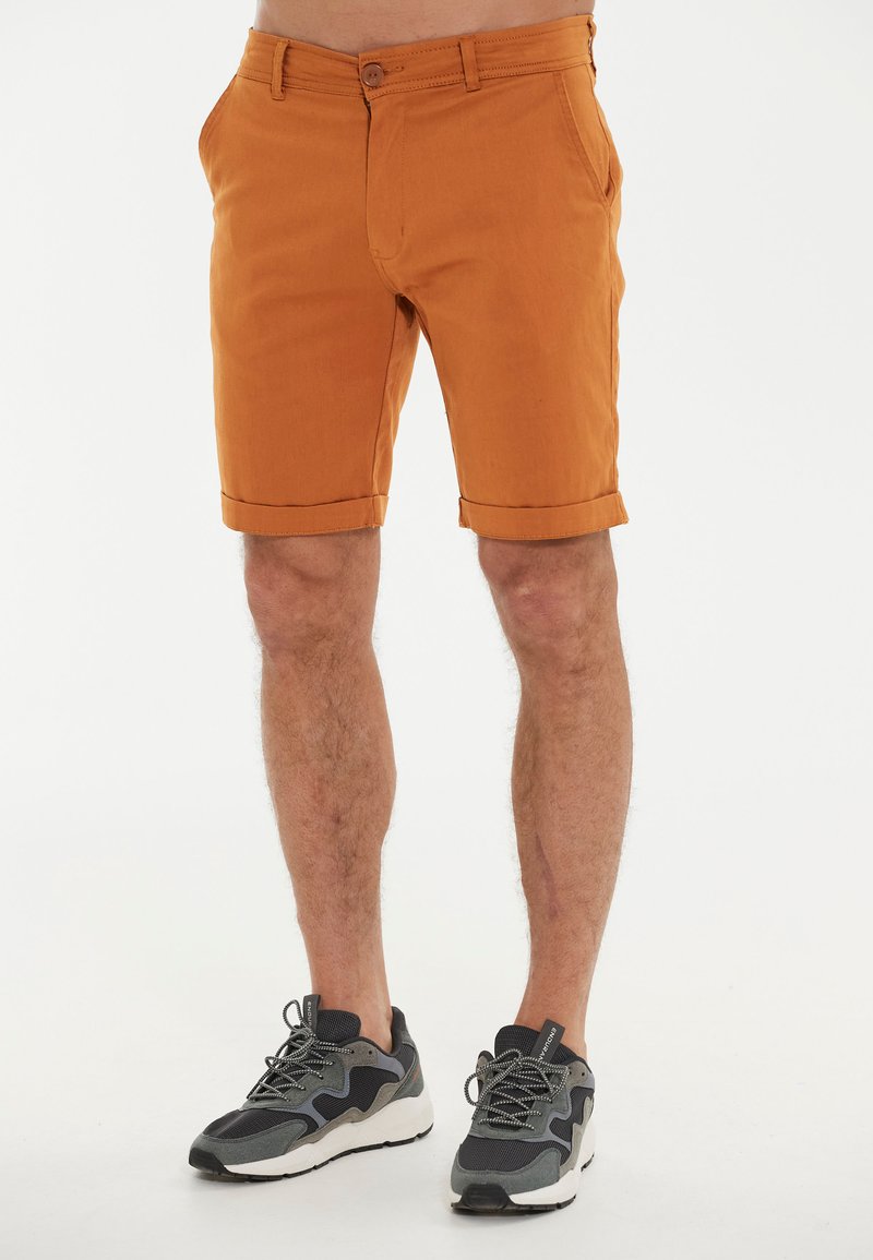Orangene Baumwollshorts mit umgeschlagenen Säumen, ausgestattet mit einem vorderen Knopfverschluss und Seitentaschen, kombiniert mit schwarzen und grauen Sportschuhen.