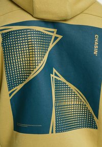 Zelena kapuca na hrbtni strani z velikimi modrimi geometrijskimi oblikami in rumenimi mrežnimi vzorci, na desni strani pa je beseda "CHASIN'" v rumenem tisku.