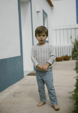 Kind in einem hellgrauen gestrickten Pullover mit strukturiertem Muster und lockerer Passform, kombiniert mit hellblauen Denim-Jeans. Barfuß auf einer Betonoberfläche.