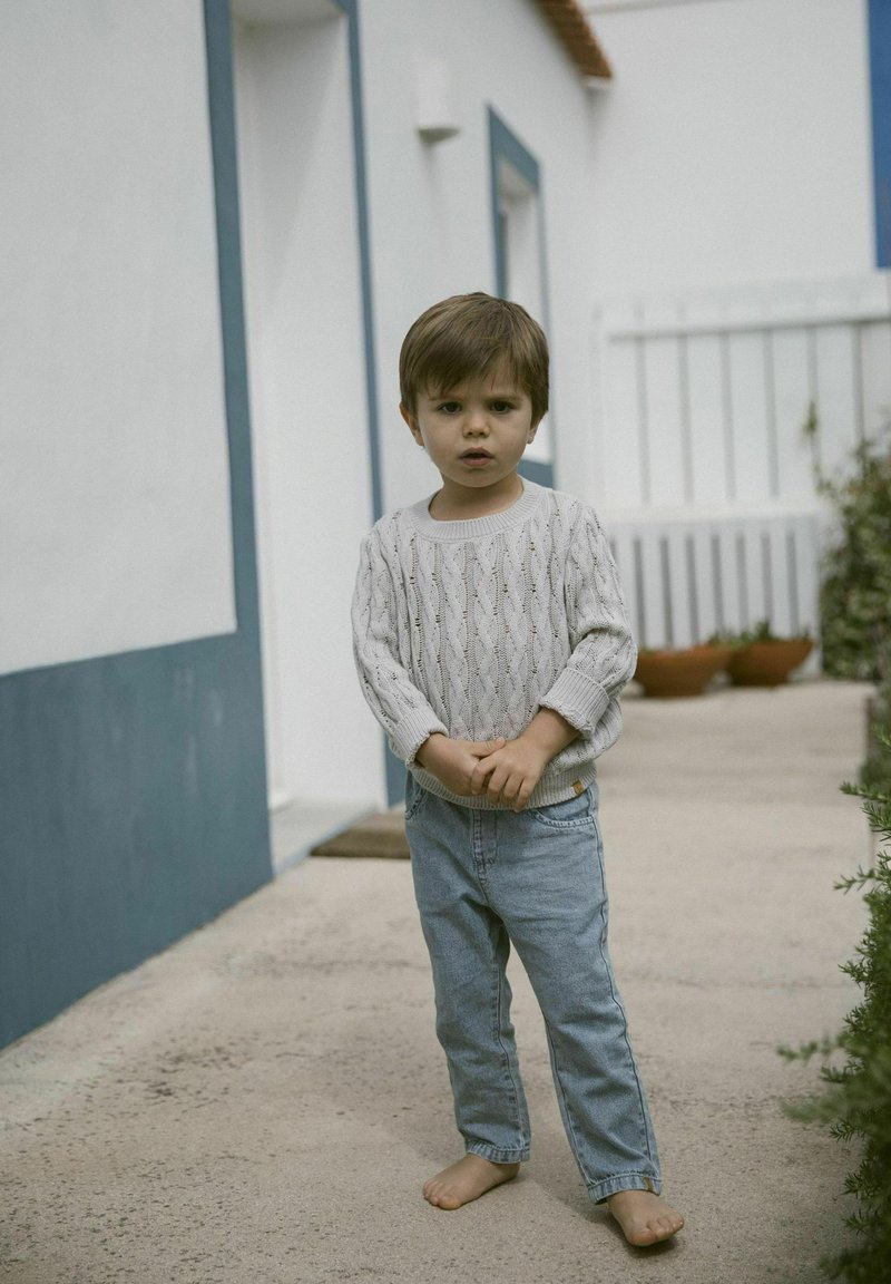 Otrok v svetlo sivi pleteni pulover s teksturo in ohlapnim krojem, v kombinaciji s svetlo modrimi jeans hlačami. Bos na betonski površini.