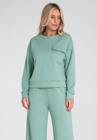 Miętowa bluza z okrągłym dekoltem i kieszenią z przodu. Wykonana z miękkiego materiału, luźna i z ściągaczami przy mankietach. Można ją skomponować z pasującymi spodniami.