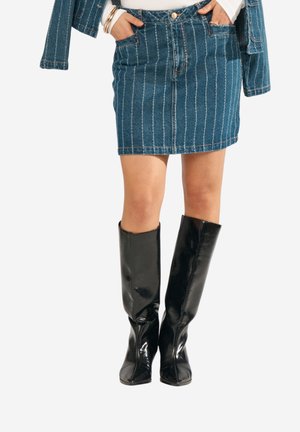 Kvinde iført en blå denimnederdel med nålestriber og matchende jakke, accessoriseret med armbånd og sorte knæhøje læderstøvler.