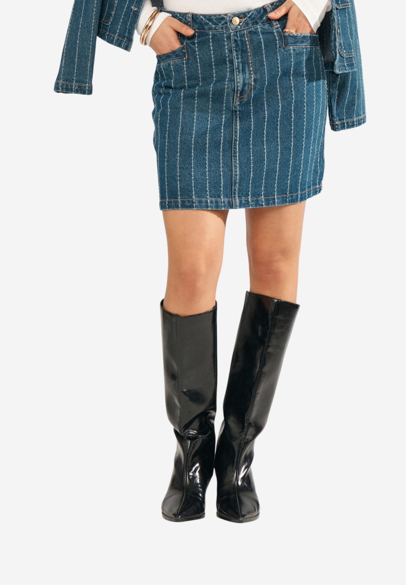 Femme portant une jupe en jean bleu à fines rayures et une veste assortie, accessoirisée avec des bracelets et des bottes en cuir noires montantes jusqu'aux genoux.