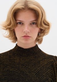 Top nero con collo alto, caratterizzato da un motivo a righe dorate texture. La modella ha capelli biondi ondulati fino alle spalle e orecchini a cerchio argentati.