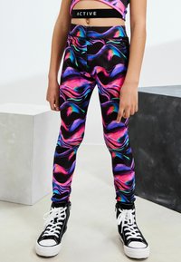 Aktive Leggings mit einer schwarzen Basis und lebhaften pinken, blauen und grünen Wirbeln. Hergestellt aus dehnbarem Material mit einer taillierten Passform, kombiniert mit schwarzen High-Top Sneakers.