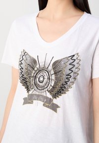 Vit T-shirt med kort ärm och V-ringning, prydd med en svart och guldig design av vingar samt en grå banner med dekorativ sömnad.