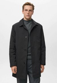 Abrigo negro con cuello con muesca, cierre de botones y mangas largas. Lleva puesto un suéter oscuro y texturizado, junto con pantalones grises. Diseño sencillo.