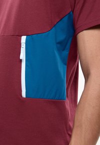 Gros plan d’un bras et d’un torse portant une chemise grenat avec une poche zippée bleue soulignée de blanc sur le côté.