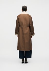 Άτομο με πλεγμένα μαλλιά που φοράει μακρύ καφέ παλτό, φαρδιά σκούρα παντελόνια και μαύρα μπότες με τακούνι, στέκεται γυρισμένο με την πλάτη προς το απλό φόντο.