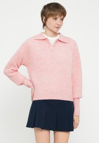 Maglione rosa lavorato a maglia con colletto, tre bottoni e polsini a costine, abbinato a una gonna blu navy plissettata. Texture liscia.