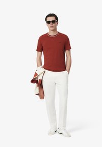T-shirt rossa con maniche corte, colletto bianco e logo, abbinata a pantaloni bianchi e una giacca bomber beige appoggiata sul braccio. Sneakers bianche.