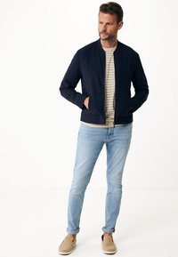 Marinblå bombarjacka, randig beige skjorta, ljusblå jeans och beige slip-in skor. Avslappnad stil med modern passform och rena linjer.