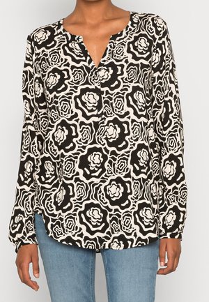 Femme portant un chemisier à manches longues noir et blanc à motif floral avec un col en V et un jean bleu, debout devant un fond uni.