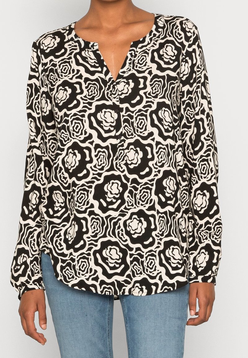 Femme portant un chemisier à manches longues noir et blanc à motif floral avec un col en V et un jean bleu, debout devant un fond uni.