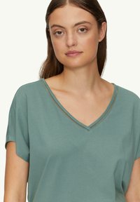 V-hals korte mouwen top in een zachte teal stof, met decoratieve stiksels langs de halslijn, een ontspannen pasvorm en een gladde textuur.