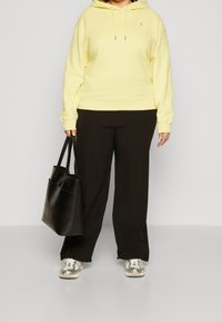Osoba w żółtej bluzie z kapturem i czarnych spodniach, trzymająca czarną torbę na ramię, w białych i szarych sneakersach, stojąca na jednolitym tle.
