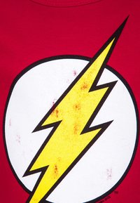 Red t-shirt featuring a large white circle with a jagged yellow lightning bolt in the center. Fabric appears cotton with slight mottling.