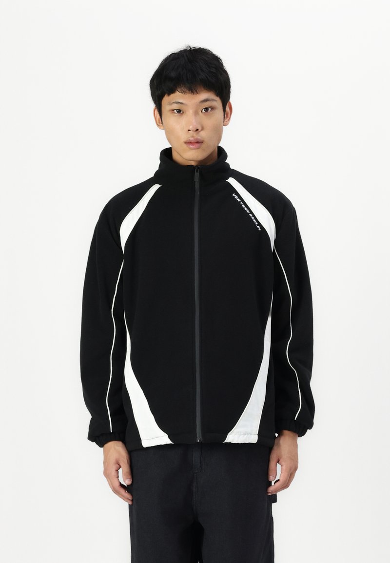 Giacca in pile nera con accenti diagonali bianchi e colletto alto. Presenta una chiusura con zip completa e polsini elasticizzati per una vestibilità aderente.