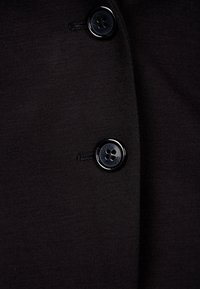 Nahaufnahme von schwarzem Stoff mit glatter Textur, zwei runden schwarzen Knöpfen und Nähdetails.