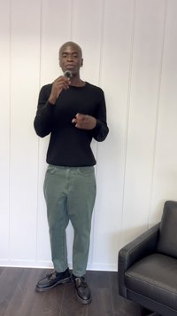 Camisa negra de manga larga, pantalones verde claro y zapatos negros con borlas. El fondo presenta una pared panelada blanca y una silla negra.