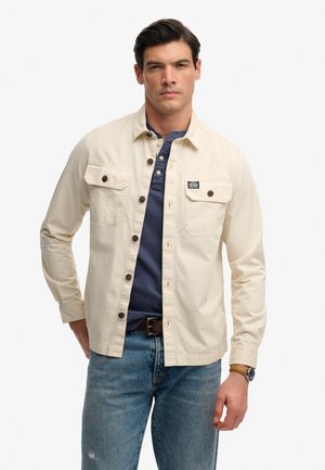 Mand iført beige skjorte over en navy henley og blå jeans, stående mod en hvid baggrund, med brun bælte og armbåndsur.