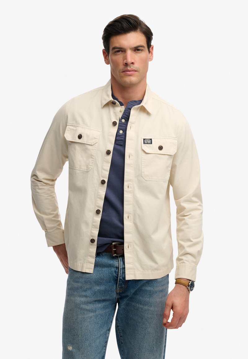 Homme portant une chemise beige boutonnée par-dessus un henley bleu marine et un jean bleu, debout devant un fond blanc, avec une ceinture marron et une montre au poignet.