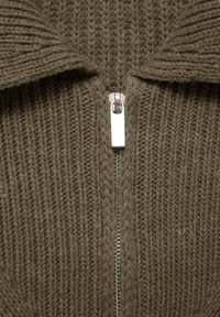 Cárdigan marrón de punto con un cuello alto y cremallera plateada. Presenta una textura acanalada que muestra líneas verticales de diferentes grosores.