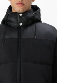 Sort polstret jakke med høj krave og snoretræk på hætten. Har branddetaljer, sølvhardware og en frontlynlås. Glat stofstruktur.