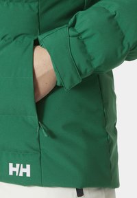 Groene geïsoleerde jas met een matte textuur, zijzak met ritssluiting en wit logo op de onderrand. Kraagdetails zijn zichtbaar.