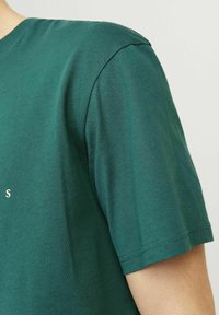 Dunkelgrünes Baumwoll-T-Shirt mit Rundhalsausschnitt, kurzen Ärmeln und einfarbigem Stoff; auf der Vorderseite befindet sich ein kleines weißes Grafikdesign.