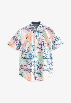 Personne portant une chemise à manches courtes avec un motif floral coloré et un pantalon gris clair, debout devant un fond vert uni.