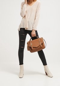 Brązowa skórzana torebka z dwoma uchwytami i metalowymi dodatkami, noszona z jasnoróżowym swetrem i czarnymi znoszonymi jeansami, zestawiona z beżowymi botkami na kostkę.