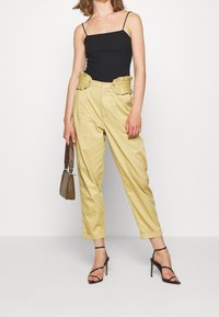 Pantalon jaune taille haute avec des accents de ceinture plissée, associé à un débardeur noir ajusté et des talons noirs à lanières. Sac à main texturé.