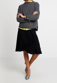 Pull en maille côtelée gris, superposé à un haut jaune, associé à une jupe noire asymétrique à volants et des escarpins noirs à motif.