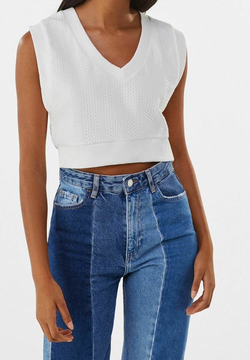 Weißes, gestricktes ärmelloses Oberteil mit V-Ausschnitt und geripptem Saum, kombiniert mit hoch taillierter zweifarbiger blauer Jeans.