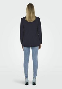 Jūras spēku zila žakete ar polsterētām pleciem, savienota ar gaiši ziliem šauriem džinsiem un bālganiem ziliem augstpapēžu apaviem. Mati taisni un plecu garumā.