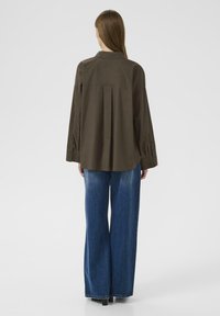 Olive green collared shirt with long sleeves, featuring pleats at the back. Paired with wide-leg blue denim jeans, showing a casual style.
