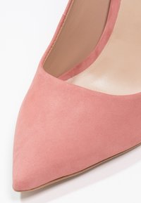 Pompe aiguille en daim rose à bout pointu, avec une texture lisse, un décolleté plongeant et un talon fin et élégant. L'intérieur est d'une teinte beige douce.