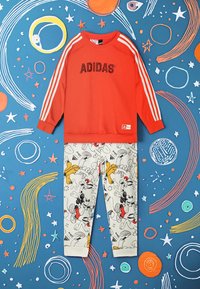 Felpa rossa con il testo "ADIDAS" e strisce bianche, abbinata a pantaloni chiari con stampe di personaggi dei cartoni animati.