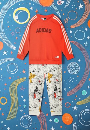 Felpa rossa con il testo "ADIDAS" e strisce bianche, abbinata a pantaloni chiari con stampe di personaggi dei cartoni animati.
