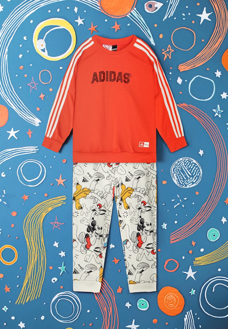 Felpa rossa con il testo "ADIDAS" e strisce bianche, abbinata a pantaloni chiari con stampe di personaggi dei cartoni animati.