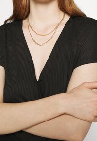 Złote, metalowe łańcuchy w warstwach opadają na szyję, kontrastując z czarną, prążkowaną bluzką z głębokim dekoltem w kształcie V i krótkimi rękawami.